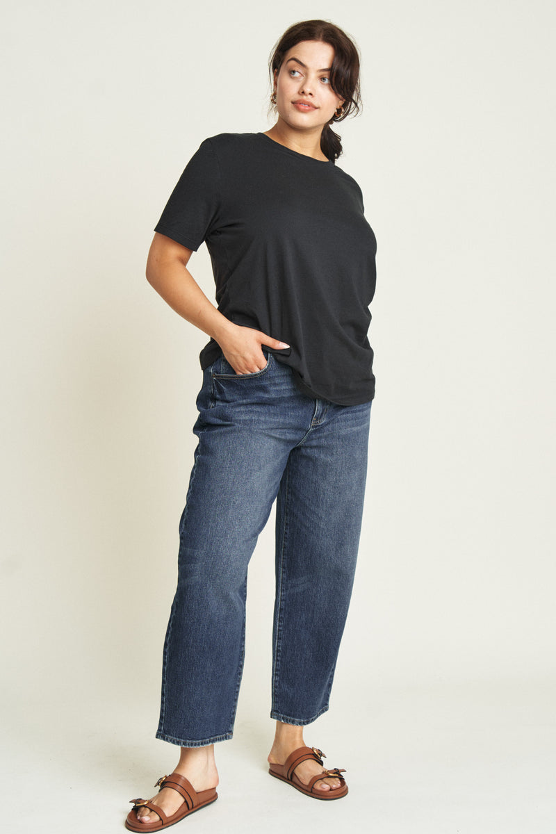 Plus-size model wearing high rise curved leg denim jeans in a dark blue wash, paired with a black t-shirt and sandals.