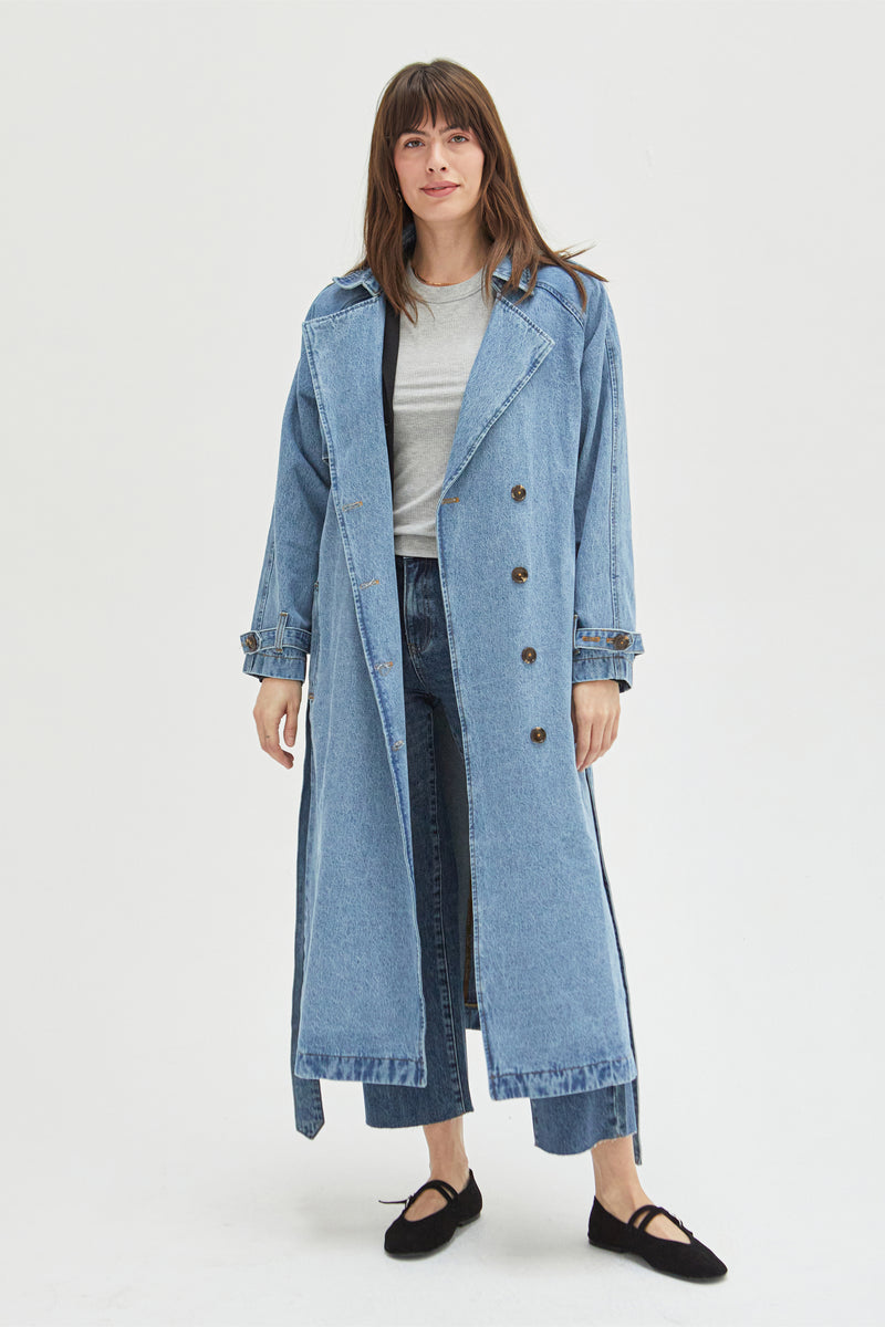 Model wearing a light wash denim trench coat styled with a gray top and light blue jeans, showcasing its longline silhouette and double-breasted button details.