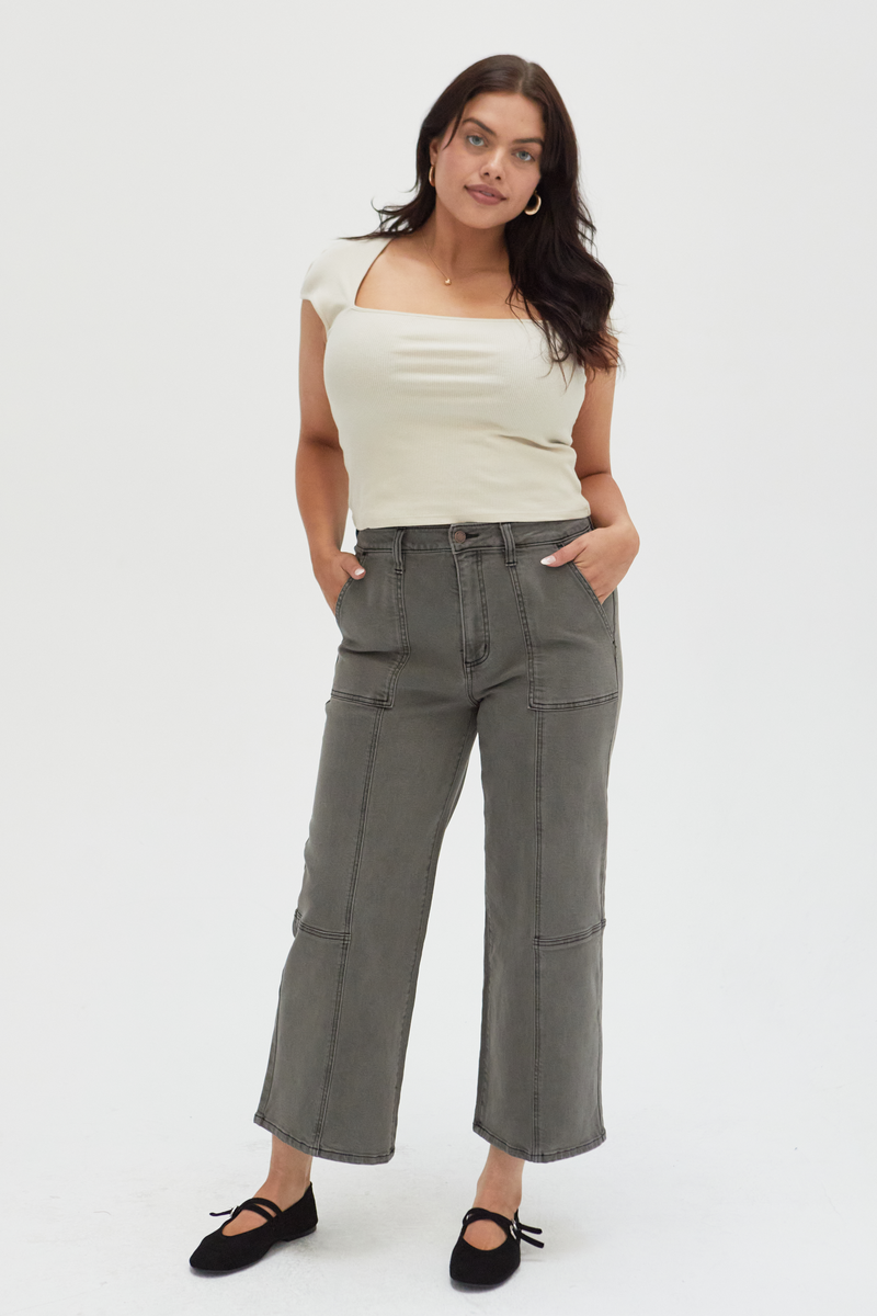 Plus-size model wearing wide-leg ankle crop jeans in an olive overdye wash, styled with a cream ribbed top and black flats, showcasing a relaxed fit and cropped hem.