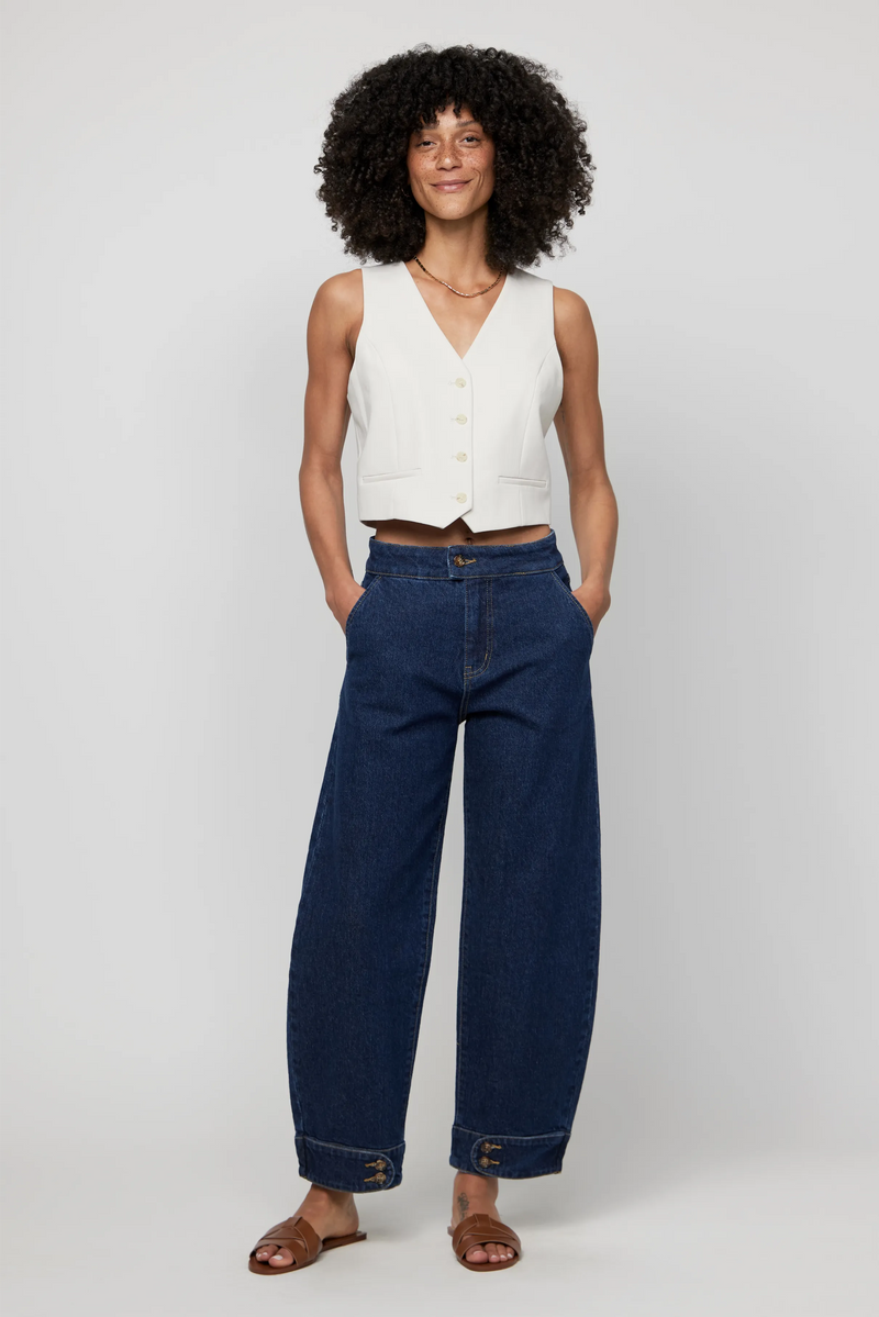Woman wearing high-rise barrel jeans in dark indigo denim, styled with a sleeveless cream vest for a modern silhouette.