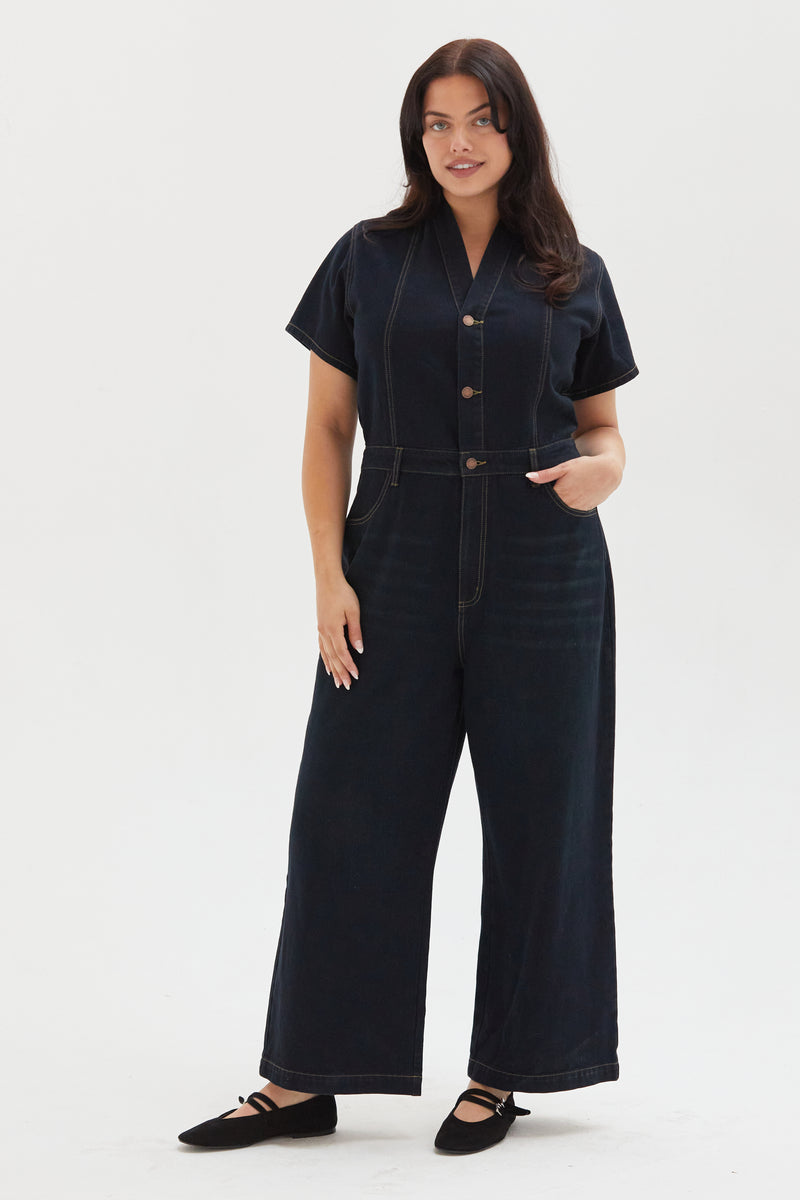 Plus-size model wearing a dark indigo one-piece denim romper with short sleeves, styled with black flats, showcasing a relaxed fit and utility-inspired design.