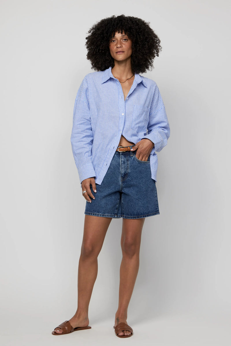 Woman in high-rise dark wash denim shorts and light blue button-down shirt, styled for casual summer wear.