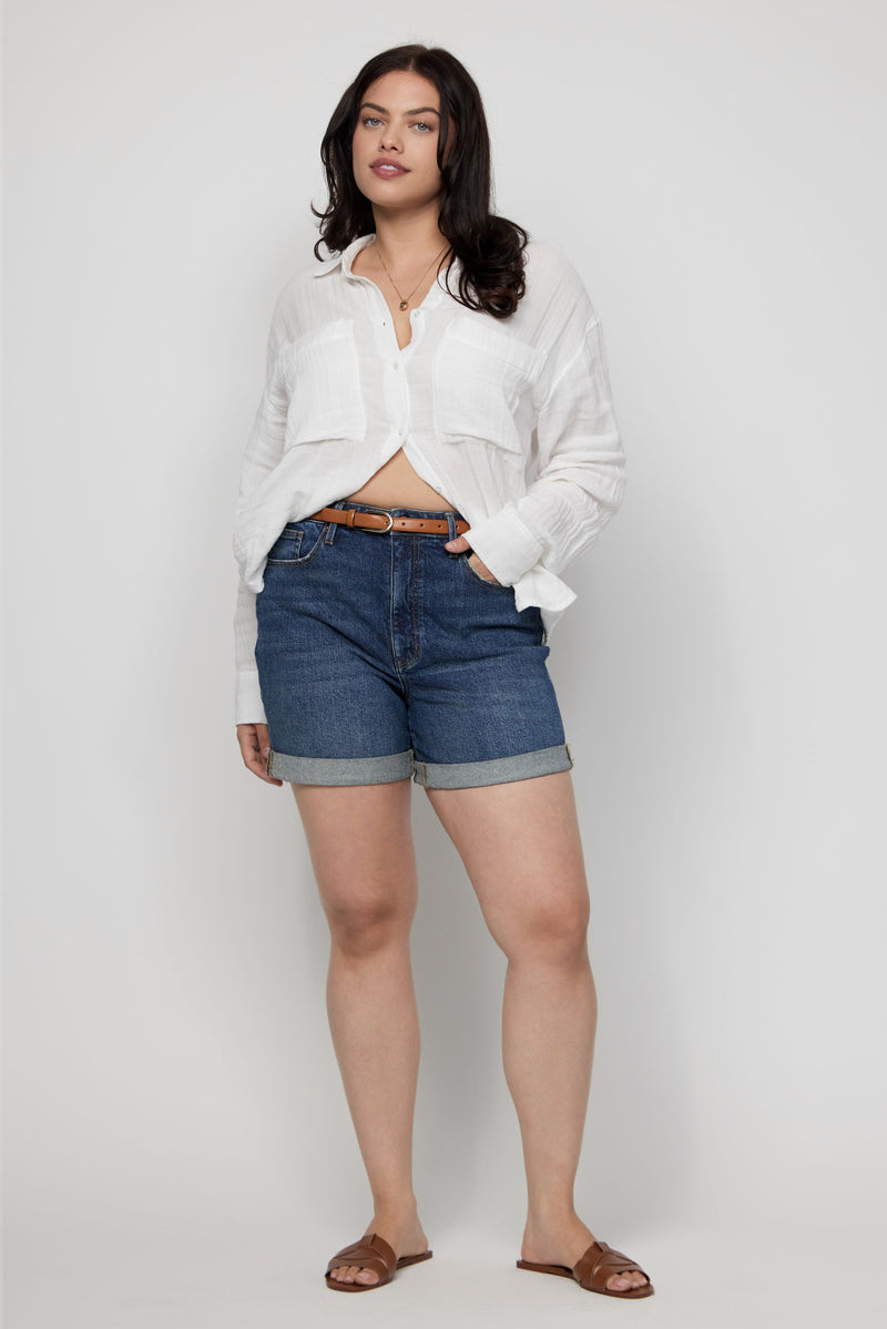 Plus-size model wearing high-rise denim shorts for women in dark indigo with a white button-down shirt.