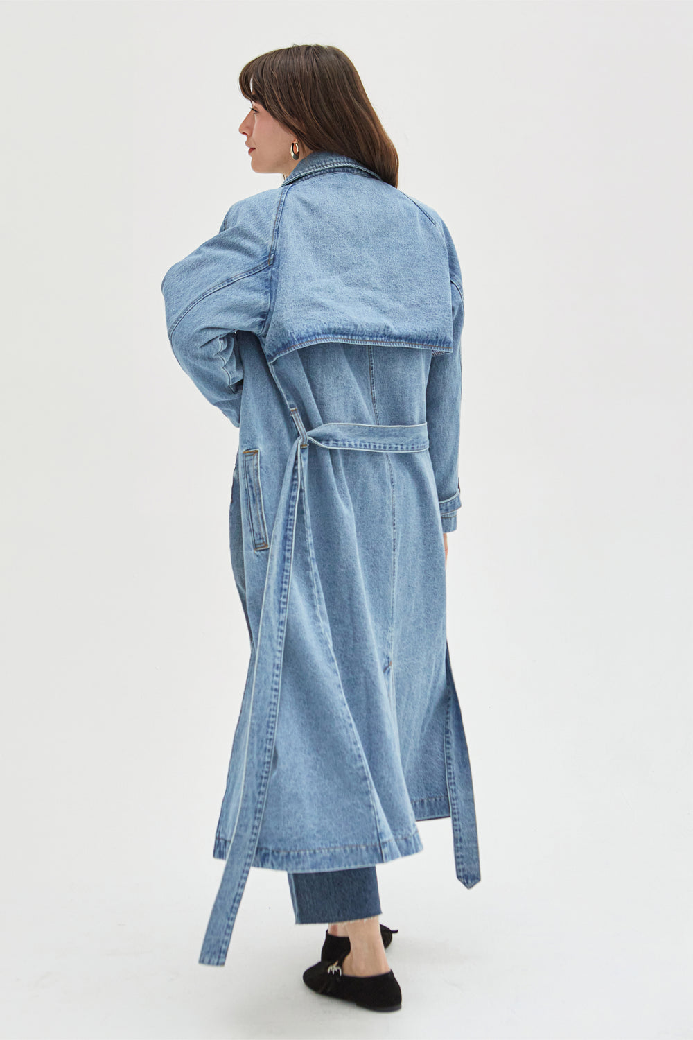 Back view of a model in a light wash denim trench coat, highlighting the tie-waist design and clean stitching details.