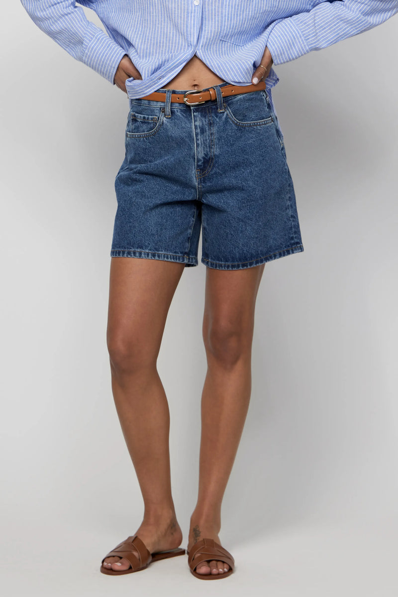 Close-up of high-rise women’s denim shorts in dark blue, styled with a brown belt and blue shirt.