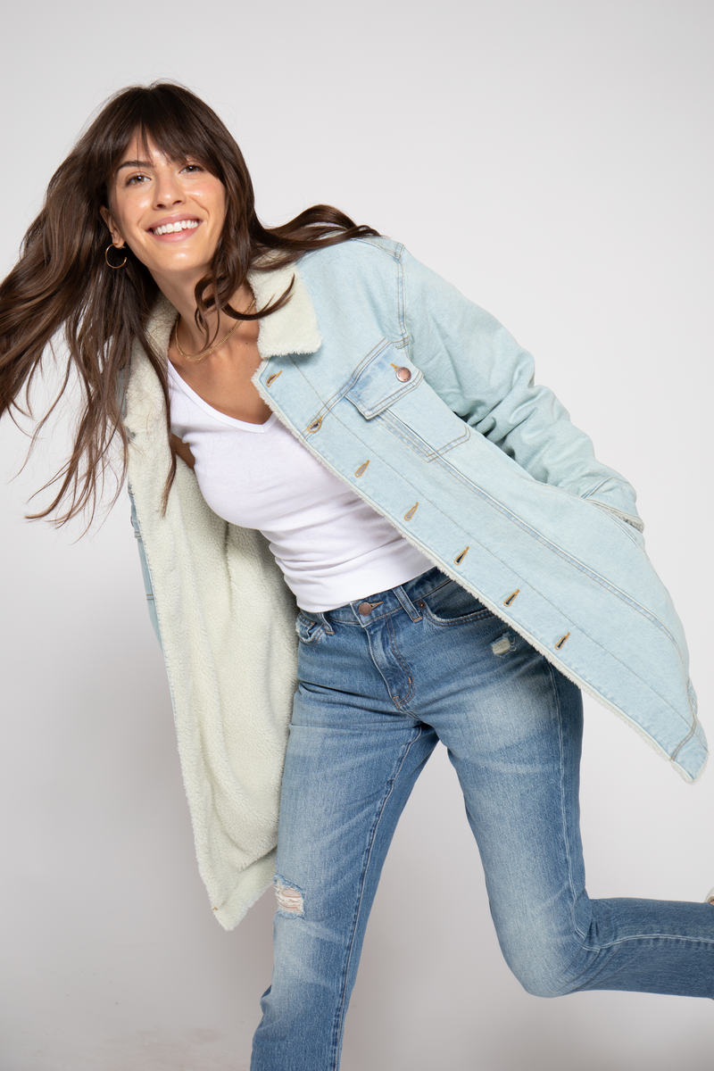 Action shot of the model smiling and opening the coat to reveal its soft sherpa lining, paired with a white top and blue jeans.