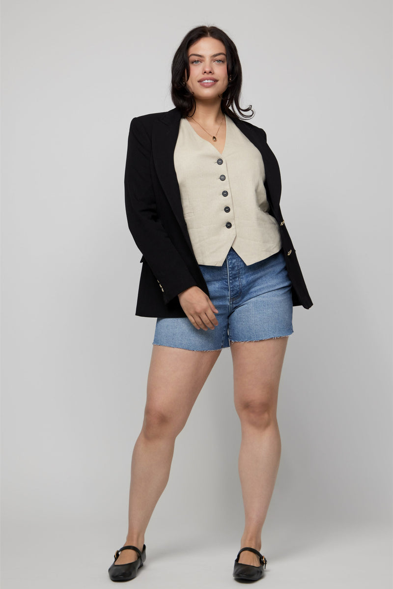Front-facing portrait of plus-size model in high-rise denim shorts, beige vest, and structured blazer.