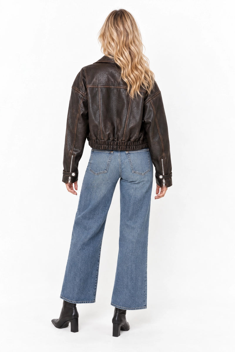 A woman viewed from the back wearing a dark brown leather jacket and wide-leg blue jeans, standing against a plain white studio background.