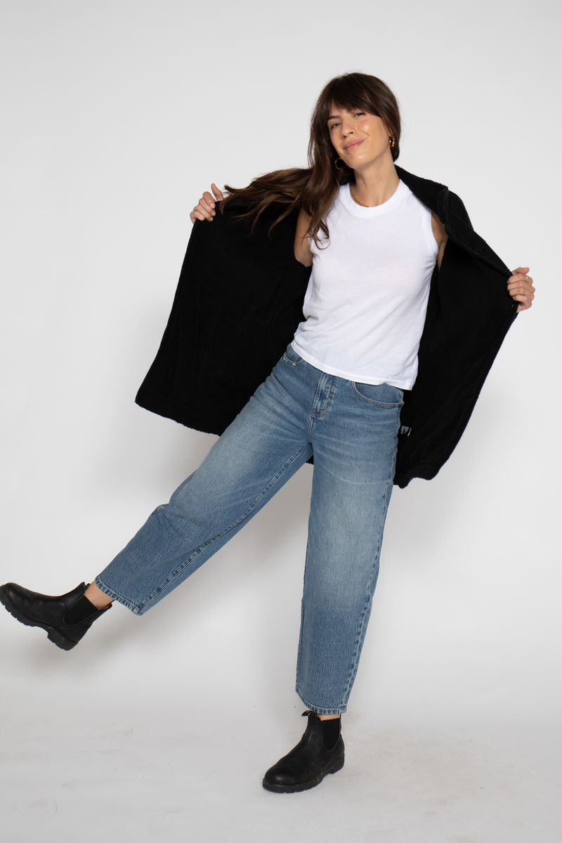 Action shot of the model opening the coat to reveal a white top underneath, paired with wide-leg jeans and boots.