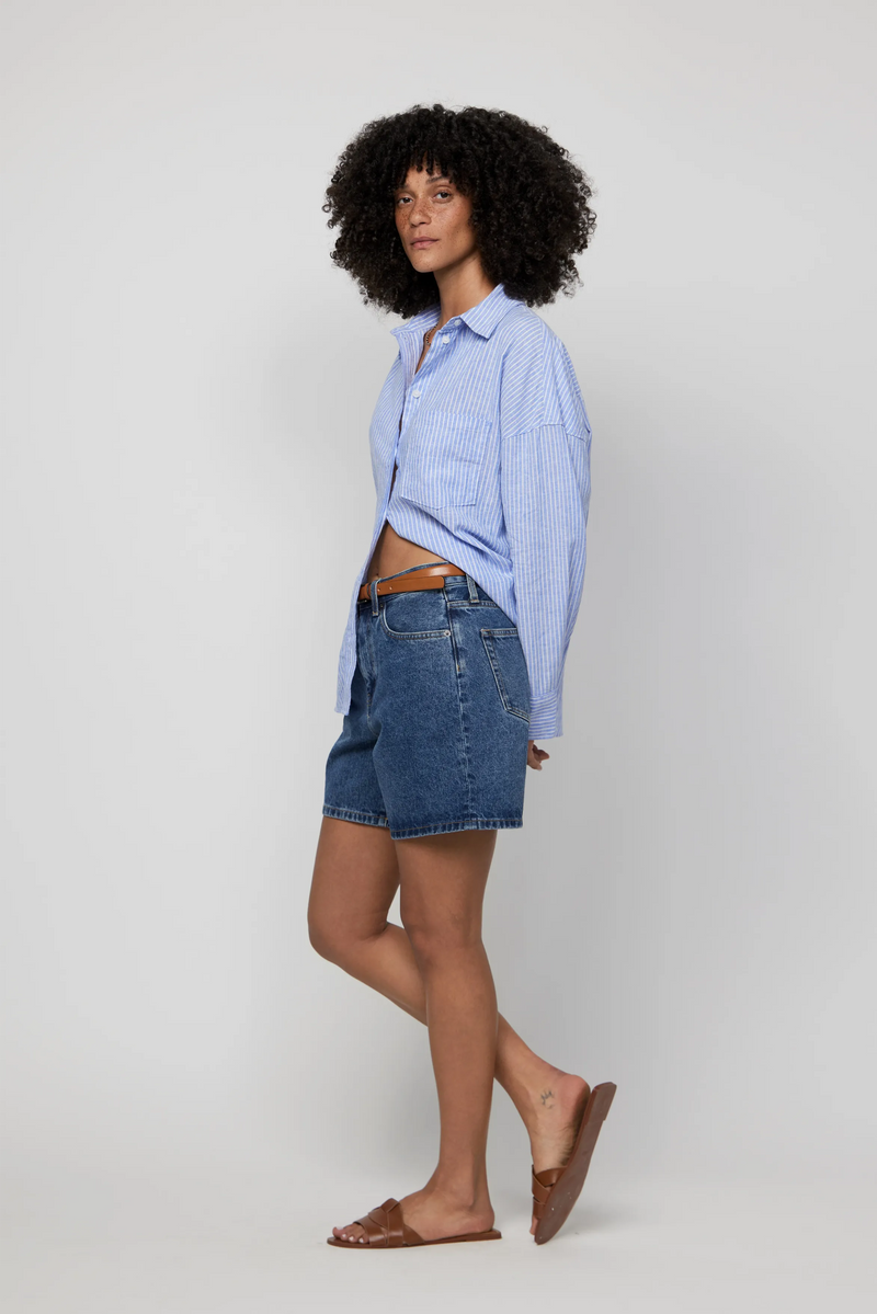 Side profile of high-rise dark wash jean shorts for women paired with leather sandals and a button-down shirt.