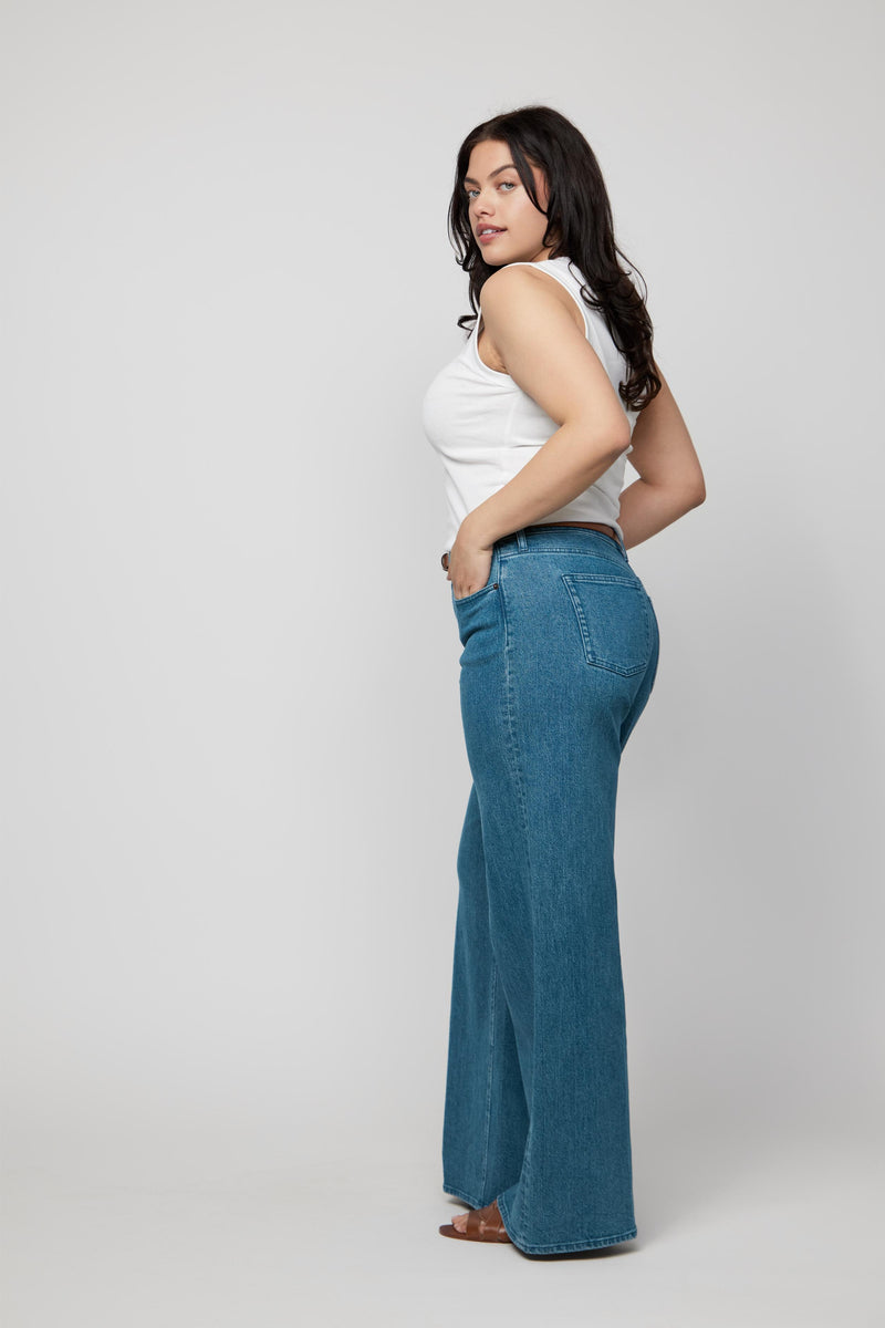 Side angle of plus-size woman in high-rise wide-leg jeans, medium wash denim paired with sandals and a white tank top.