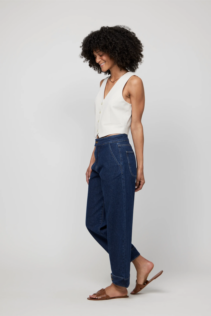 Side angle of model in dark wash high-rise barrel jeans with subtle taper at the ankle, paired with sandals and vest.