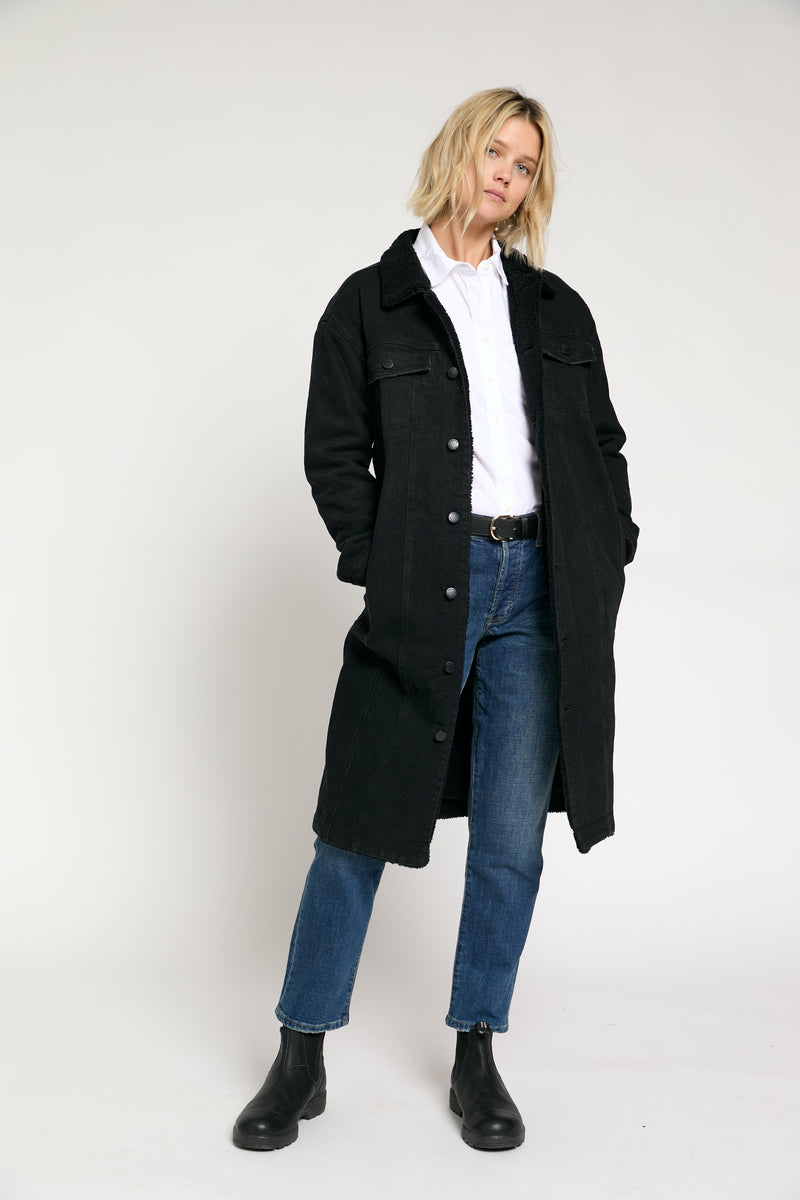 Front view of a model wearing a black sherpa-lined long trench coat, styled with jeans and a white top for a casual look.