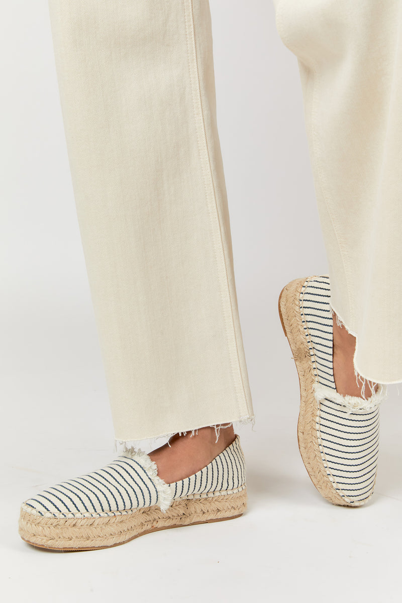 Close-up of frayed hem on ecru denim jeans, paired with striped espadrilles, highlighting the detailed design.