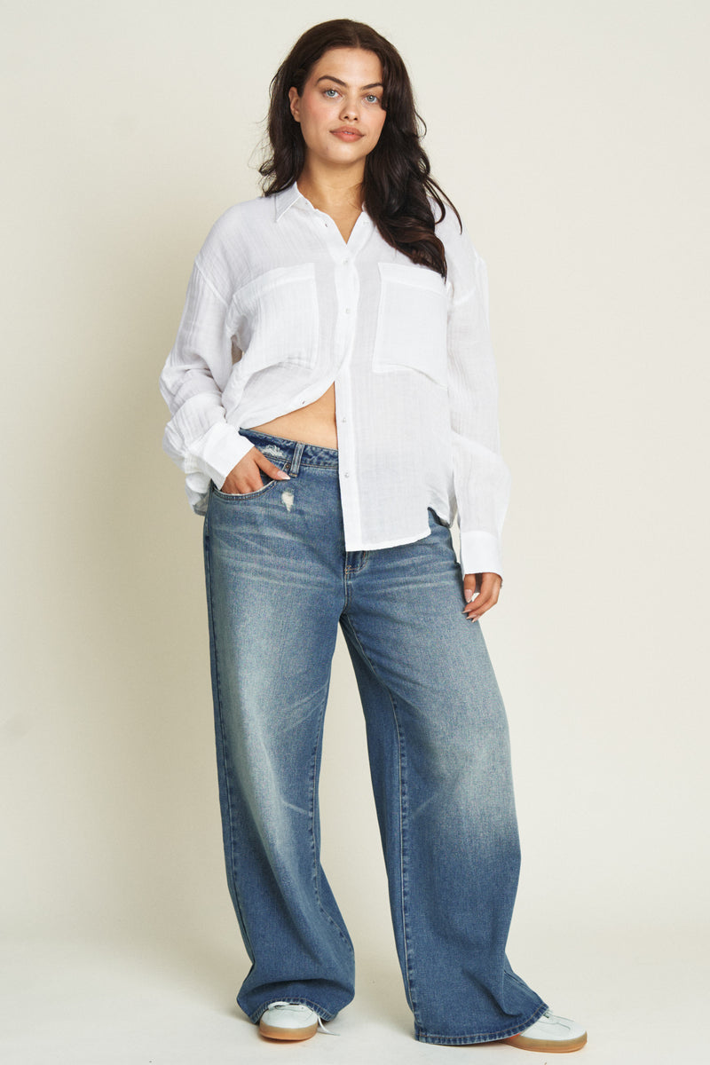 Curvy model wearing mid-rise wide-leg jeans in a medium blue wash, styled with a white cardigan and cream loafers, showcasing a flattering fit through the waist and hips with a flowing wide-leg design.
