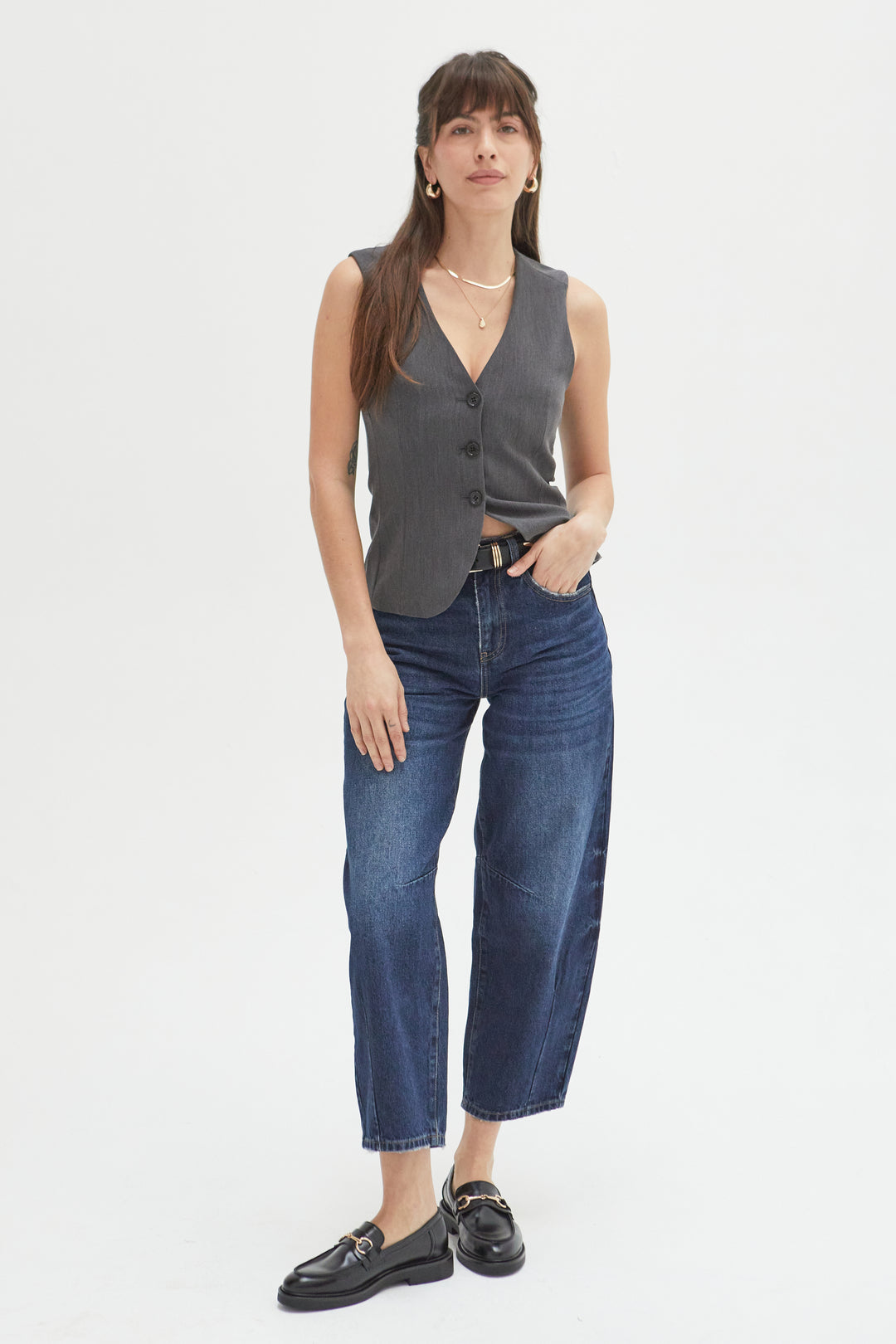 Model wearing high-rise barrel jeans in a dark indigo wash, styled with a charcoal gray tank and black loafers, showcasing a curved leg and cropped ankle design.
