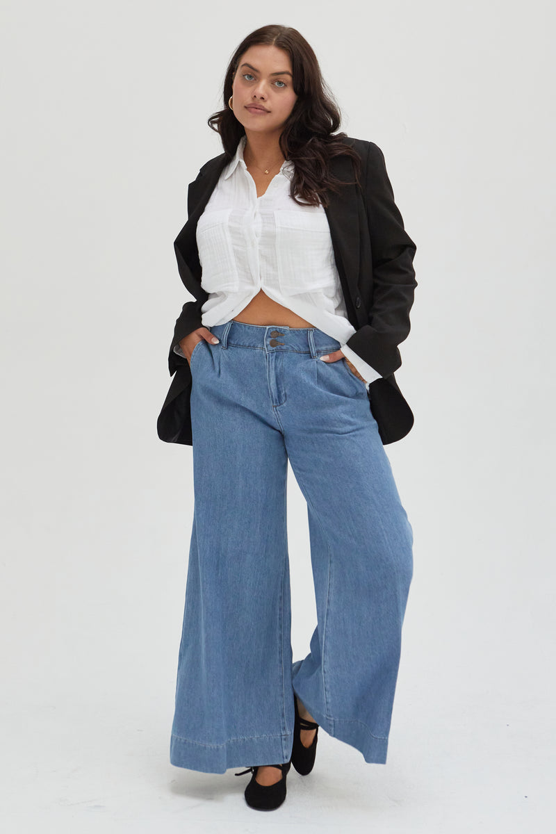 Curvy model wearing lighter blue pleated trouser low-rise wide-leg pants, styled with a white top and sneakers, showcasing a relaxed fit and elegant wide-leg silhouette.
