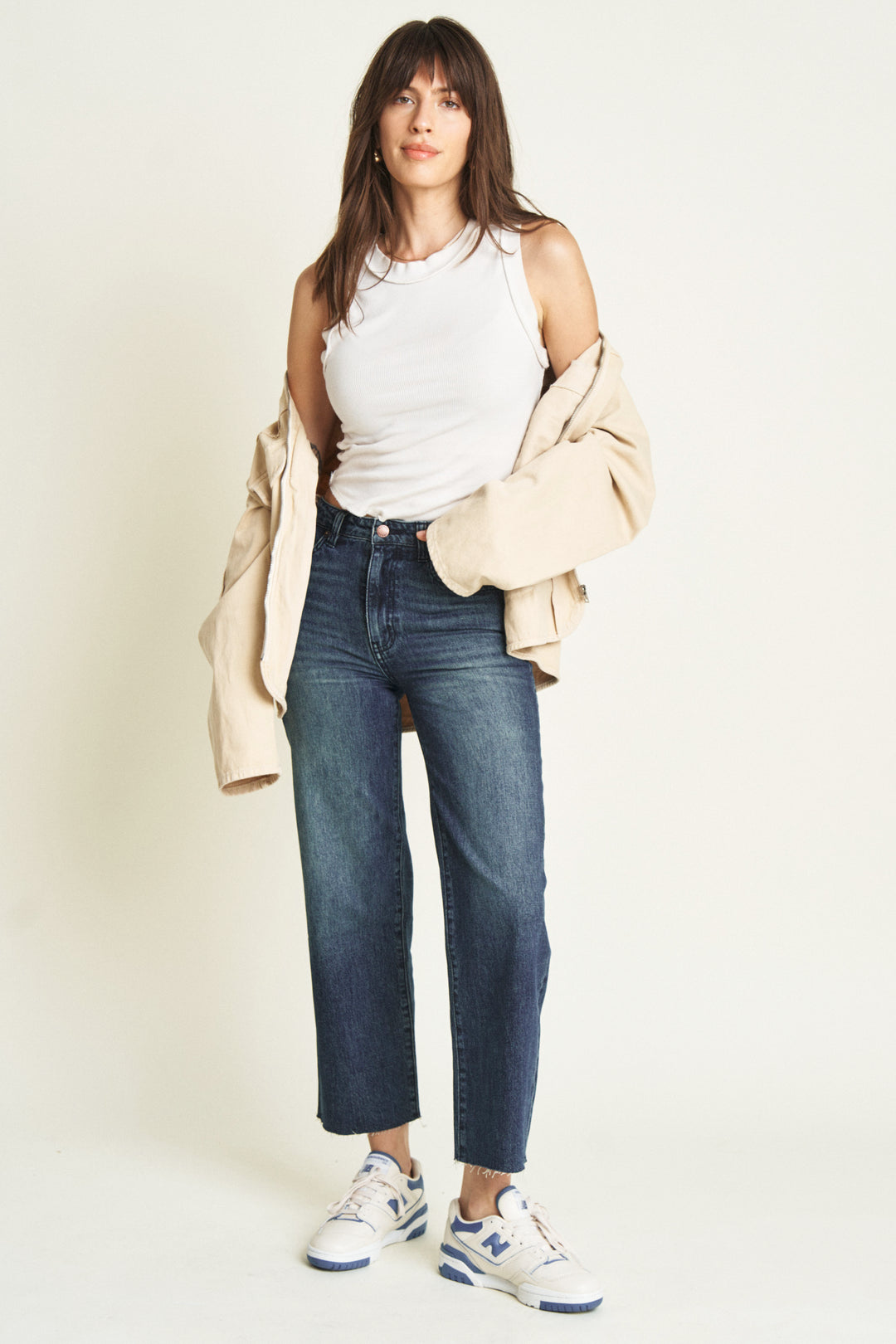 Model wearing ankle crop jeans in a dark blue wash, styled with a white top, beige jacket, and blue sneakers, showcasing a slim fit and cropped length.
