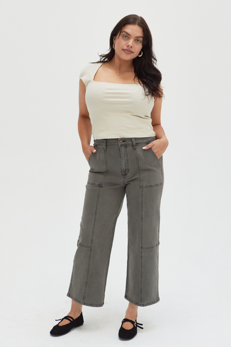 Plus-size model wearing wide-leg ankle crop jeans in an olive overdye wash, styled with a cream ribbed top and black flats, showcasing a relaxed fit and cropped hem.
