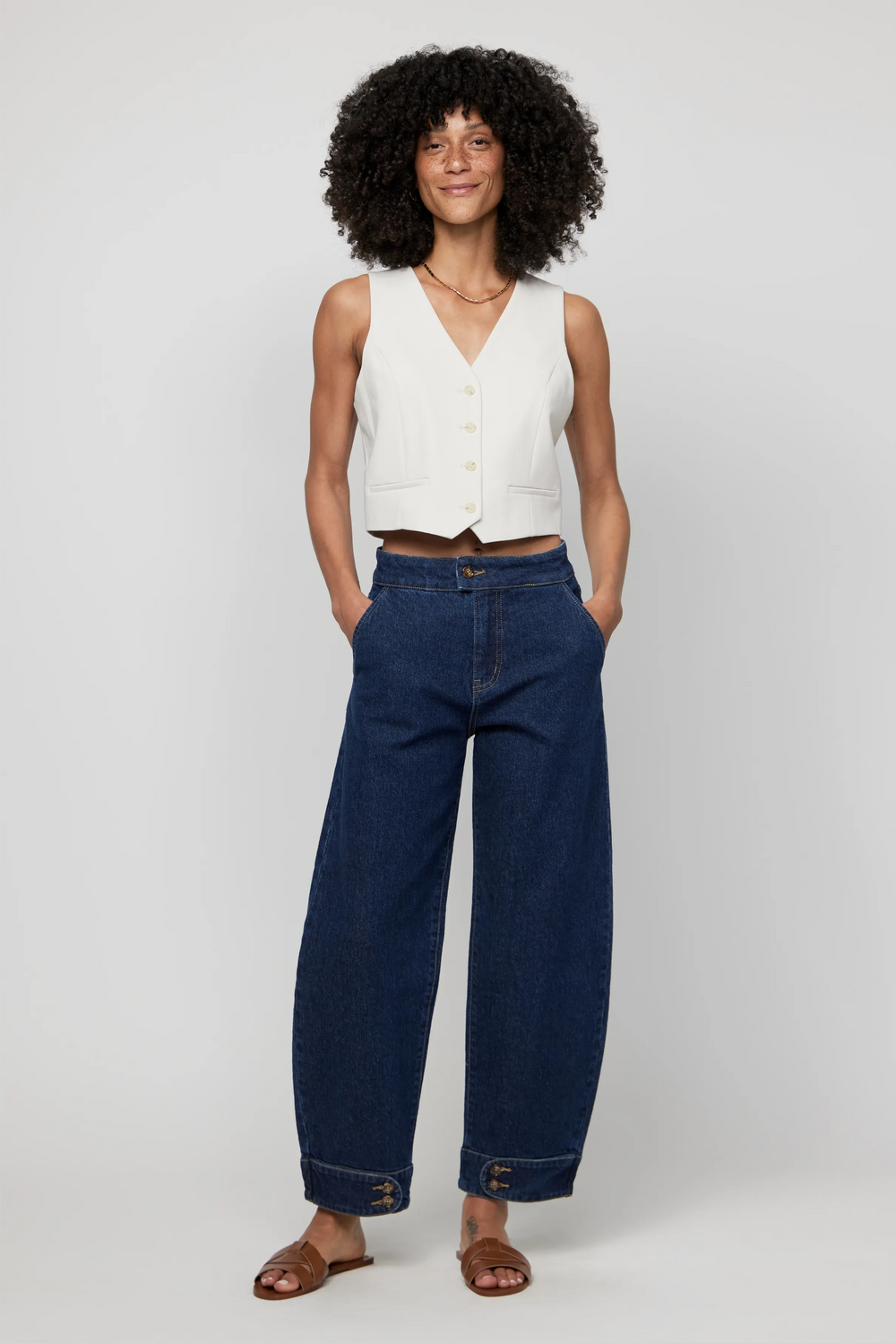Woman wearing high-rise barrel jeans in dark indigo denim, styled with a sleeveless cream vest for a modern silhouette.
