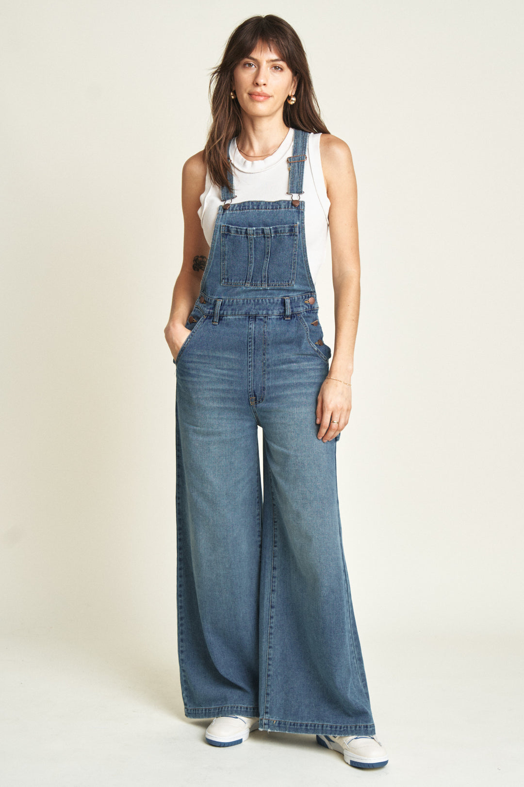 Model wearing denim overalls in a medium blue wash with a wide-leg design, styled with a white sleeveless top and white sneakers, showcasing a relaxed and casual look.