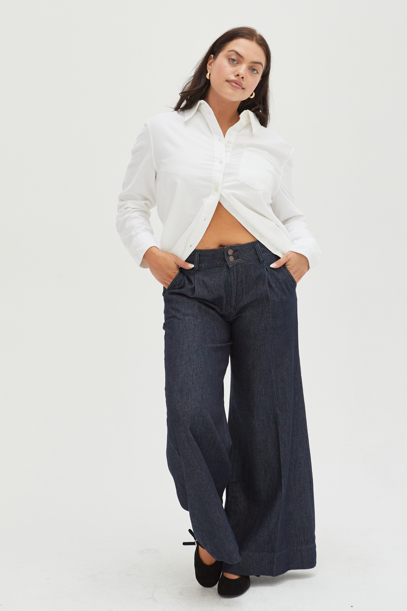 Curvy model wearing dark wash pleated trouser low-rise wide-leg pants, styled with a black blazer and white sneakers, showcasing a relaxed fit and flattering wide-leg design.
