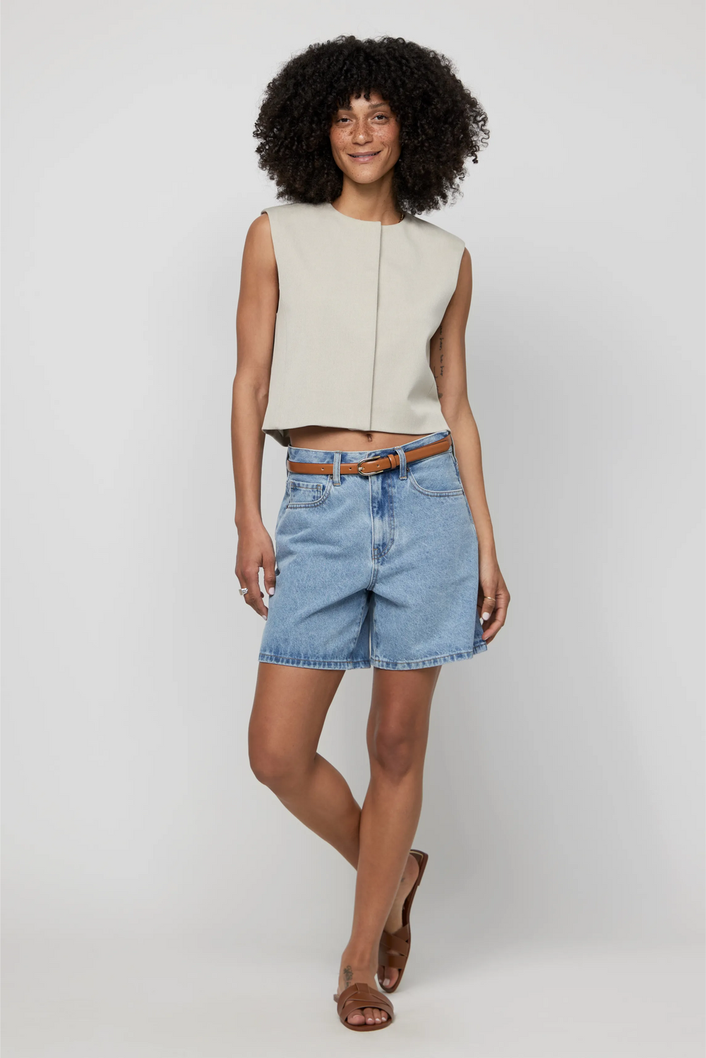 Woman in high-rise light wash denim shorts and a sleeveless beige top styled with sandals.