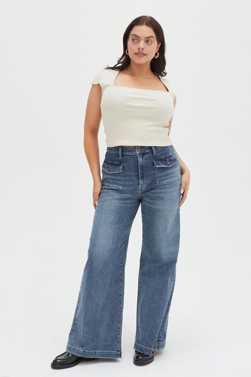 Curvy model wearing high-rise wide-leg jeans in a medium blue wash, styled with a tan cable-knit sweater and beige boots, showcasing a relaxed fit and full-length wide-leg silhouette.