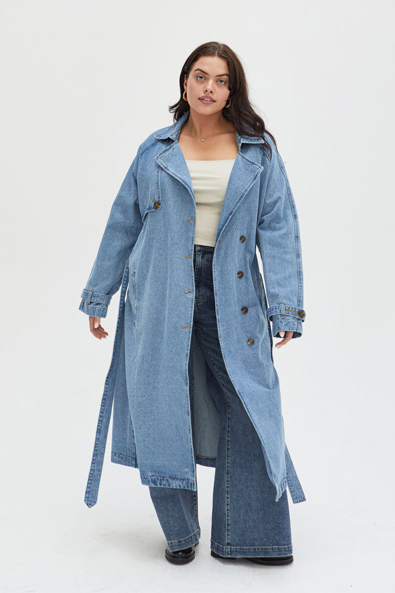 Curvy model wearing a light wash denim trench coat styled with a gray top and light blue jeans, showcasing its longline silhouette and double-breasted button details.
