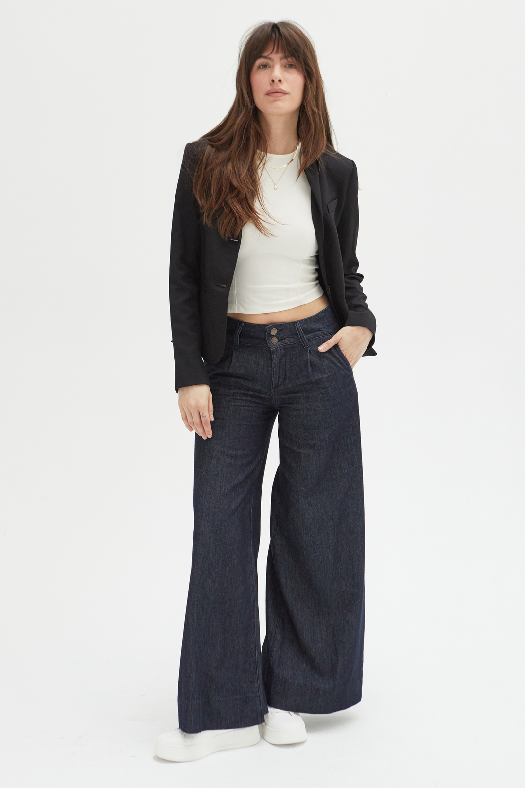 Model wearing dark wash pleated trouser low-rise wide-leg pants, styled with a black blazer and white sneakers, showcasing a relaxed fit and flowing silhouette.
