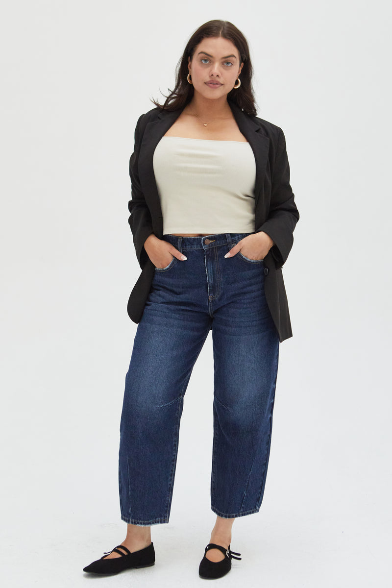 Plus-size model wearing high-rise barrel jeans in a dark indigo wash, styled with a cream top, black blazer, and black flats, showcasing a curved leg and cropped ankle fit.
