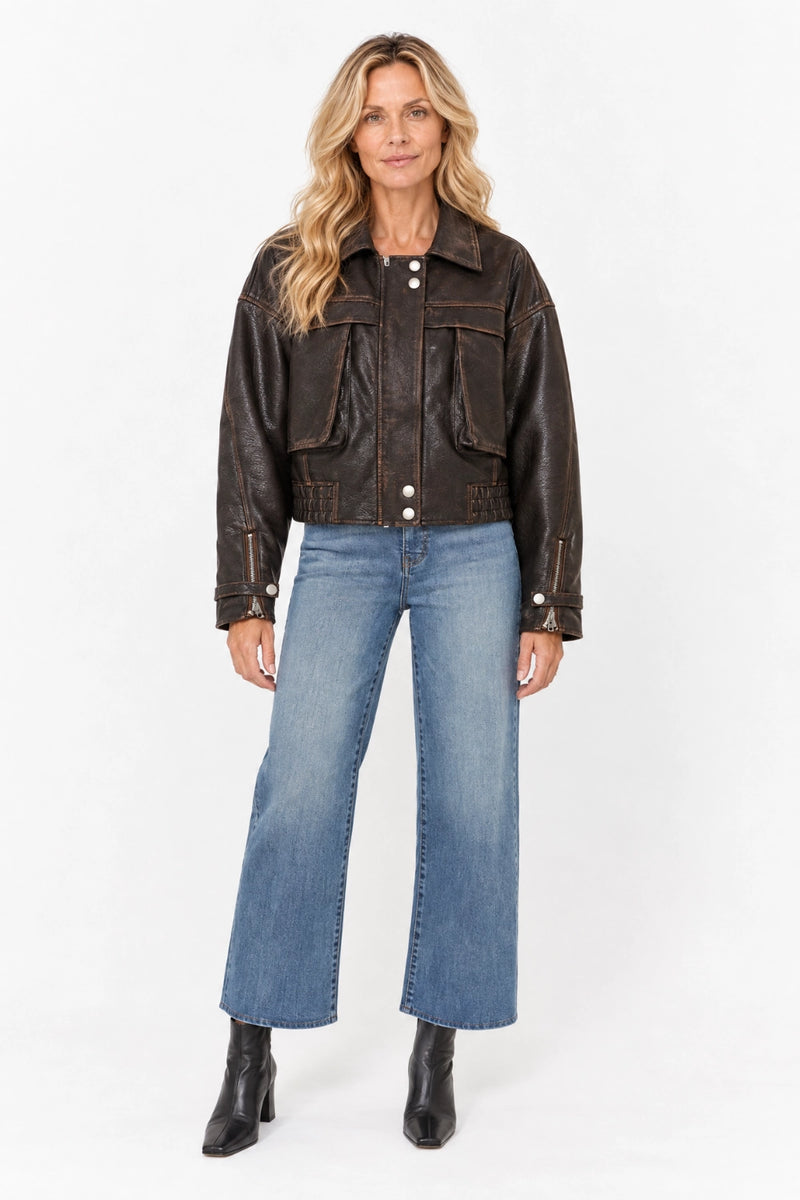 A woman modeling a dark brown leather jacket with front flap pockets, paired with blue wide-leg jeans and black ankle boots, against a plain white studio background.