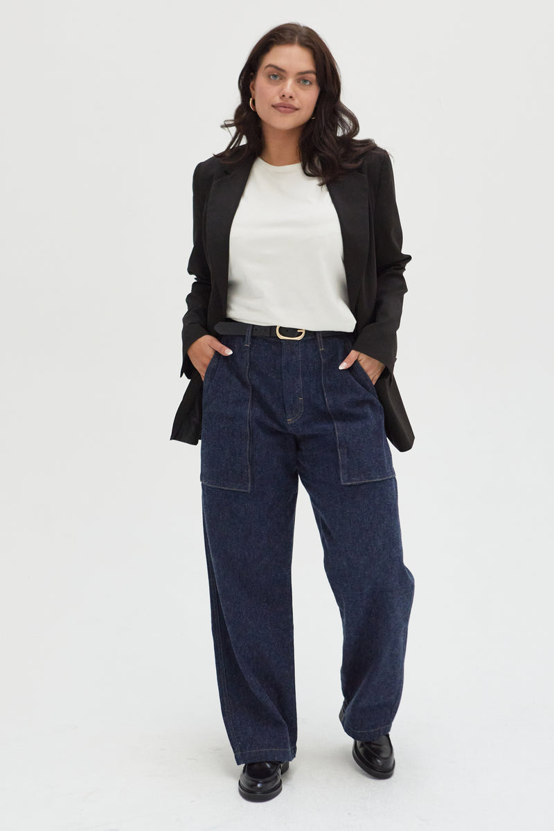 Plus-size model wearing trouser jeans in a dark indigo wash, styled with a white top, black blazer, and black loafers, showcasing a relaxed fit and wide leg with front pocket detailing.
