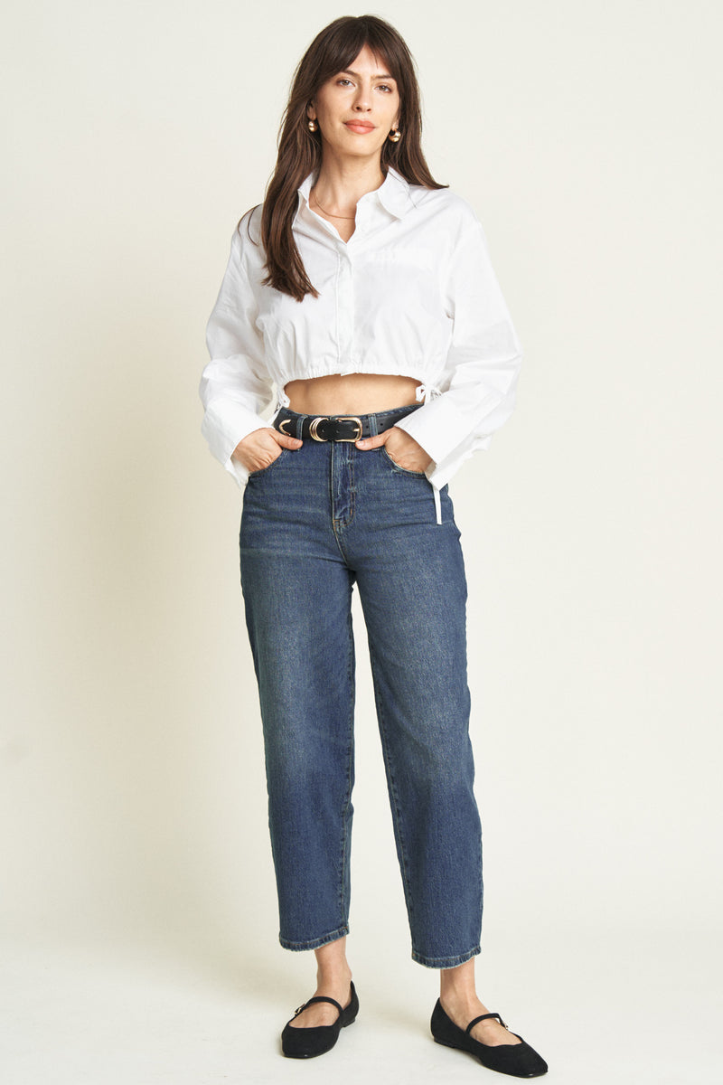 Front view of model wearing high rise barrel jeans in a classic blue wash, paired with a white cropped blouse and black flats.
