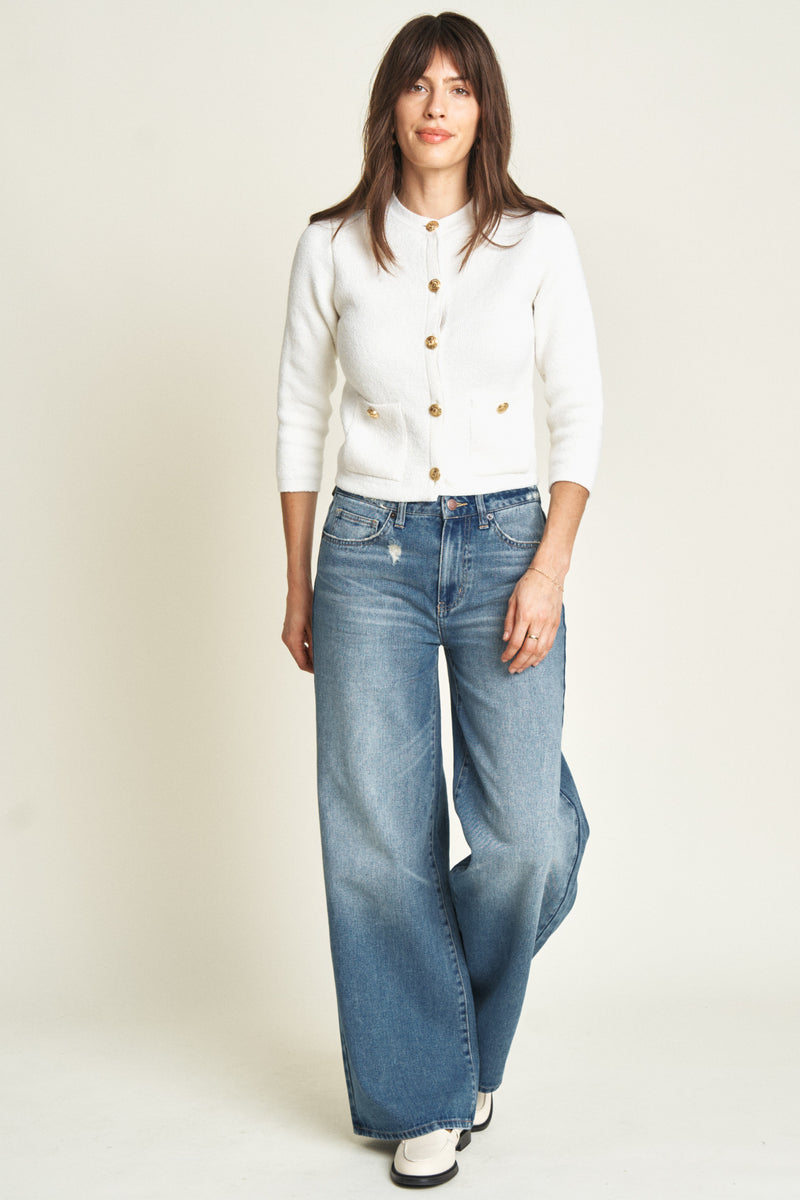 Model wearing mid-rise wide-leg jeans in a medium blue wash, styled with a white cardigan and cream loafers, showcasing a relaxed fit and flowing wide-leg silhouette.
