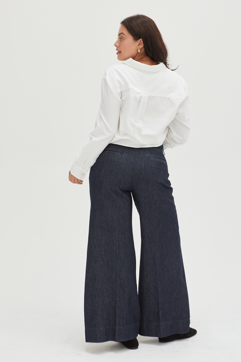 Back view of a curvy model in dark wash low-rise wide-leg pants, emphasizing the pleated detailing and tailored fit through the hips.
