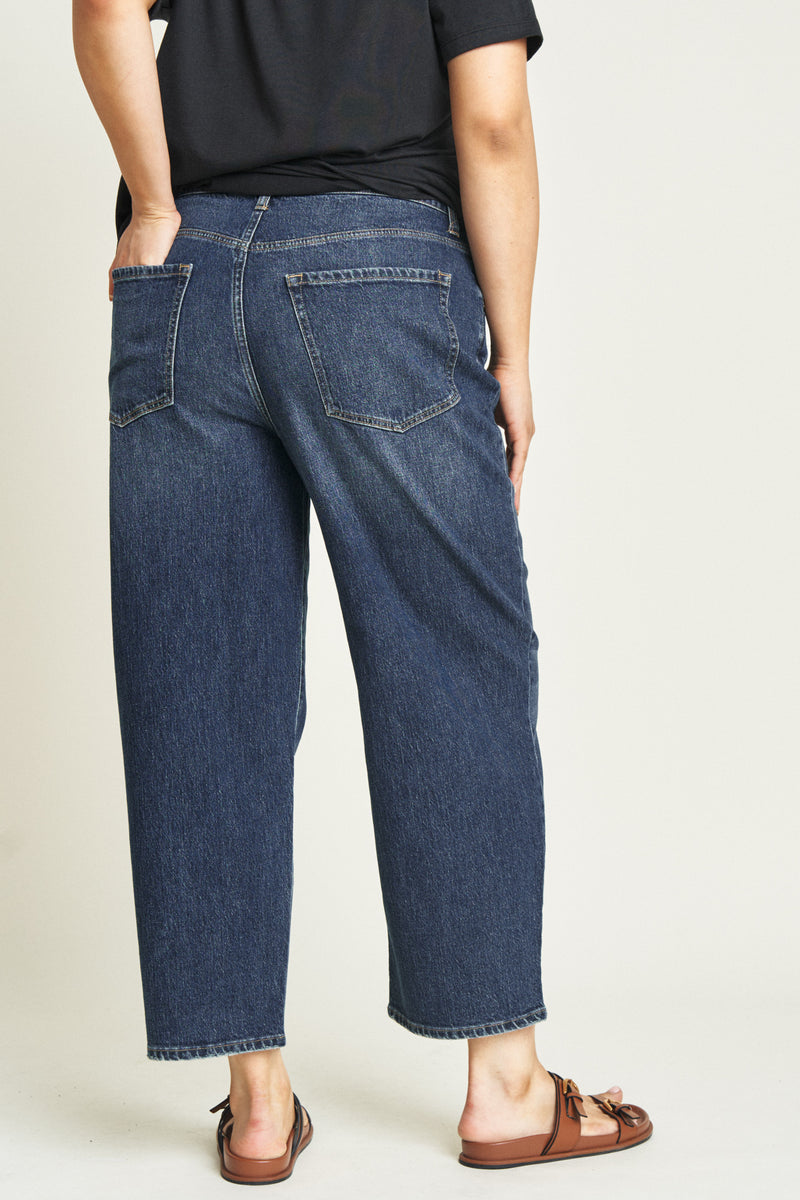 Back view of plus-size model in curved leg jeans, showing the relaxed fit and clean dark blue wash, paired with sandals.
