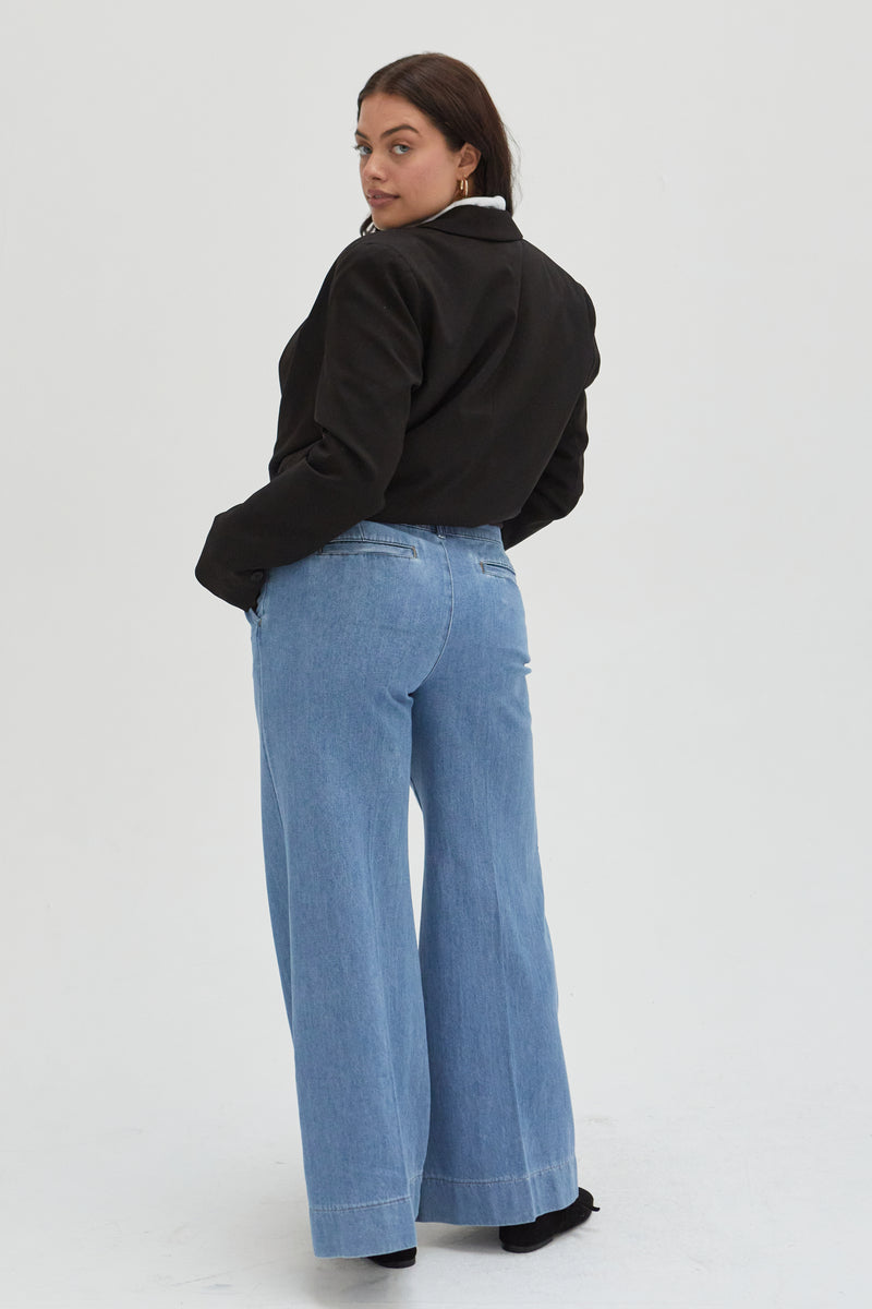 Back view of a curvy model in lighter blue low-rise wide-leg pants, emphasizing the tailored pleats and flattering fit through the hips.
