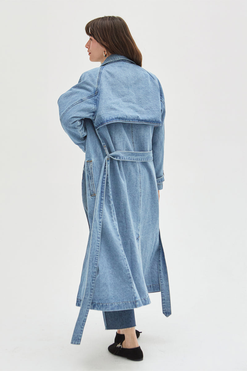 Back view of a model in a light wash denim trench coat, highlighting the tie-waist design and clean stitching details.
