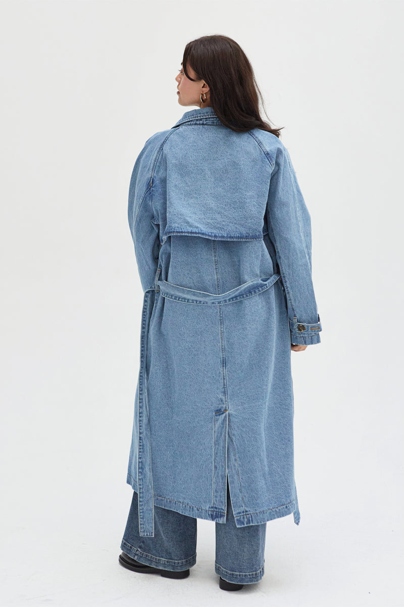 Back view of a curvy model in a light wash denim trench coat, emphasizing the tie-waist design and flattering fit through the shoulders.
