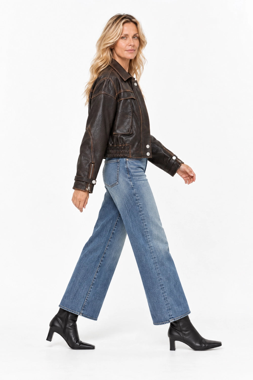 A woman modeling a dark brown cropped leather jacket, blue wide-leg jeans, and black heeled ankle boots against a plain white studio background.