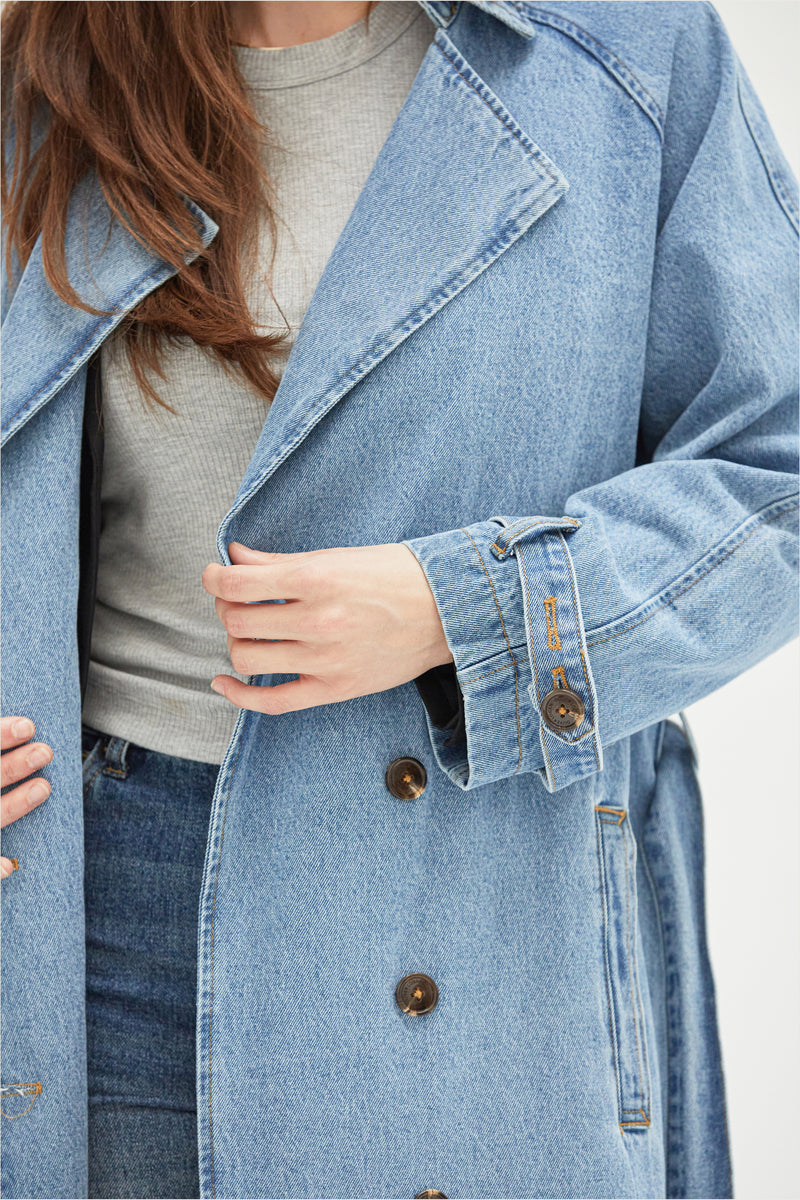 Close-up of the light wash denim trench coat, emphasizing the structured lapels, adjustable sleeve straps, and durable fabric.
