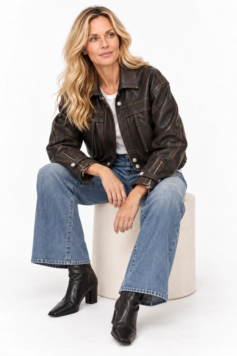 A woman sits on a white cylindrical pedestal wearing a dark brown leather jacket, blue flared jeans, and black ankle boots against a plain white studio background.
