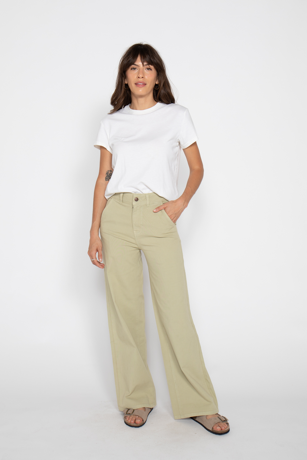Woman standing in lightweight high-waisted flare pants, hands in pockets, casual white t-shirt, on a plain background.
