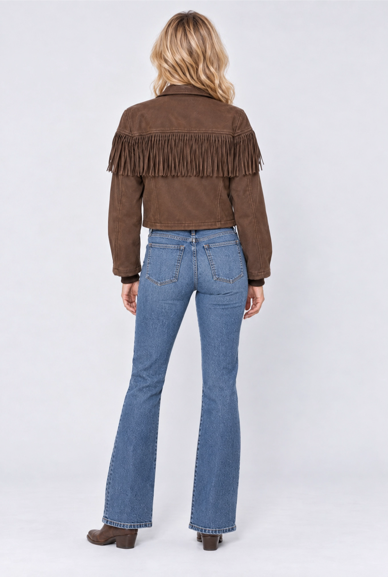 A woman viewed from the back wearing a brown suede fringe jacket and blue flare jeans, standing against a light gray studio background.
