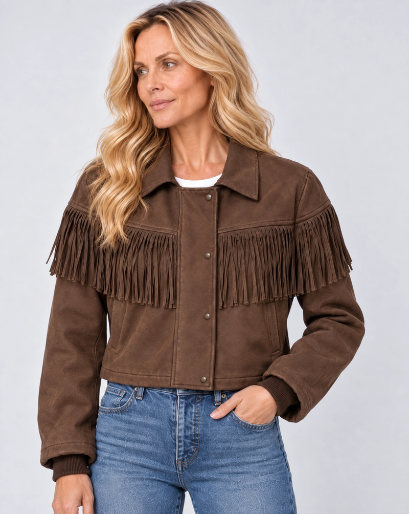 A woman wearing a brown suede fringe jacket and blue jeans, photographed against a light gray studio background.