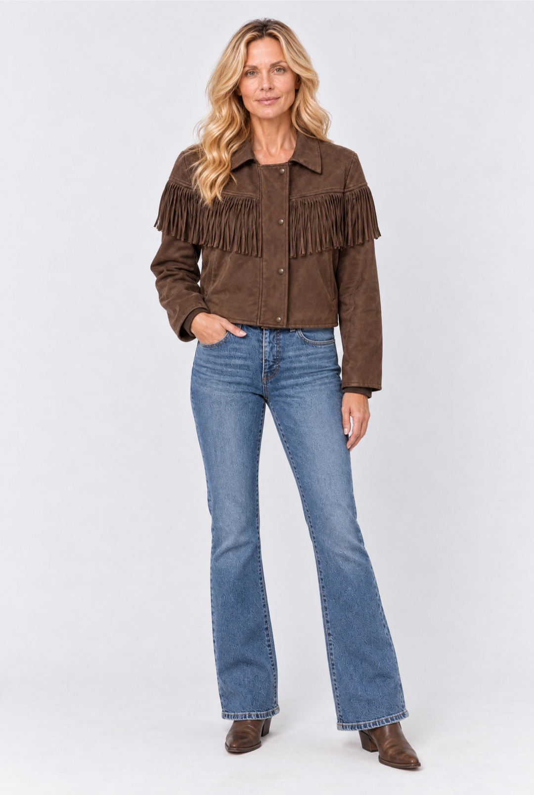 A woman modeling a brown suede fringe jacket with blue flare jeans and brown boots, standing against a light gray studio background.