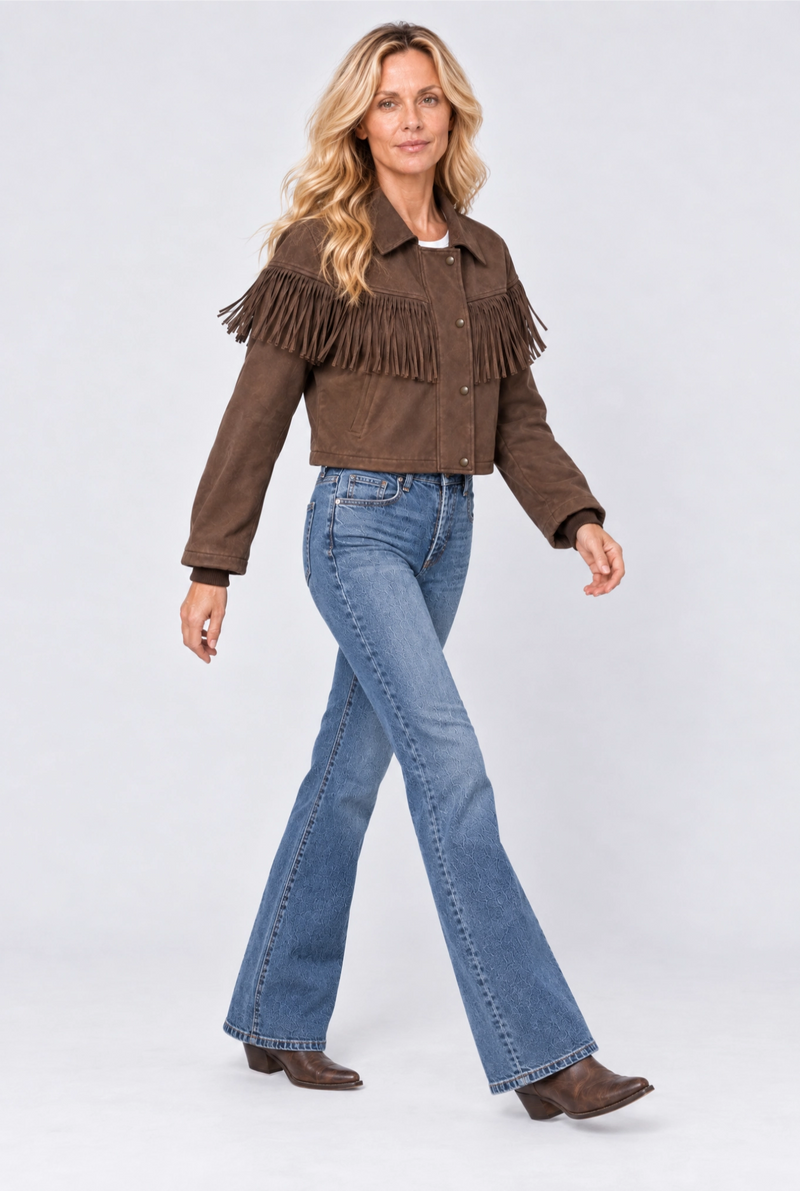 A woman modeling a brown suede fringe jacket with blue flare jeans and brown boots, standing against a light gray studio background.