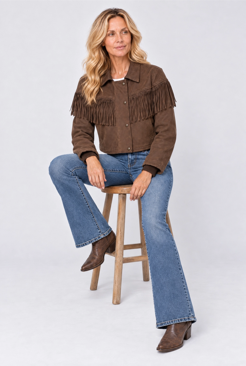 A woman sits on a wooden stool wearing a brown suede fringed jacket, blue jeans, and brown ankle boots against a light gray studio background.