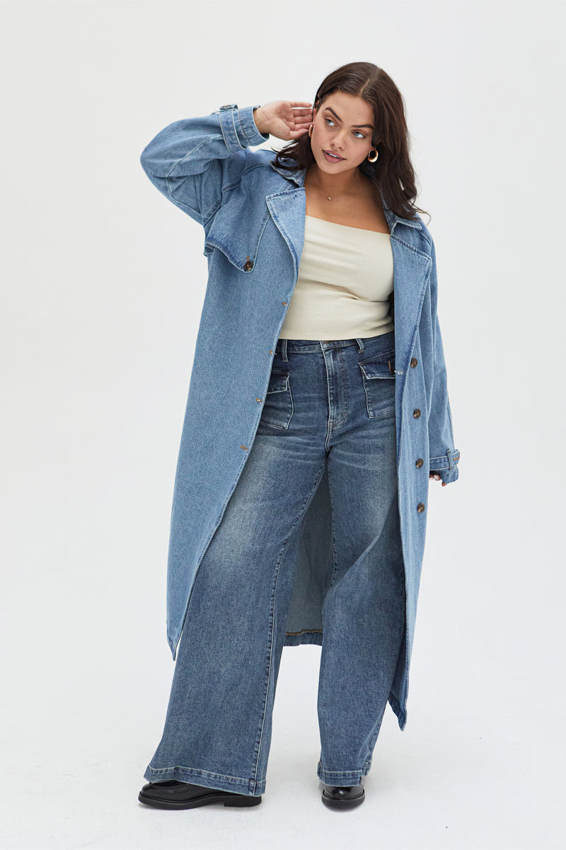 Curvy model moving in a light wash denim trench coat, demonstrating its relaxed fit and timeless appeal for any occasion.